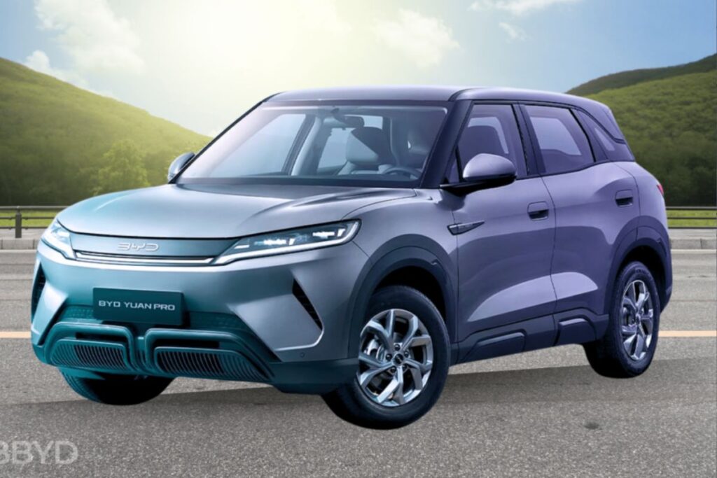 Um SUV compacto BYD Yuan Pro cinza-azulado estacionado em uma estrada com montanhas e céu ensolarado ao fundo. O carro possui o logotipo "BYD" na parte dianteira e "BYD YUAN PRO" na placa de identificação.